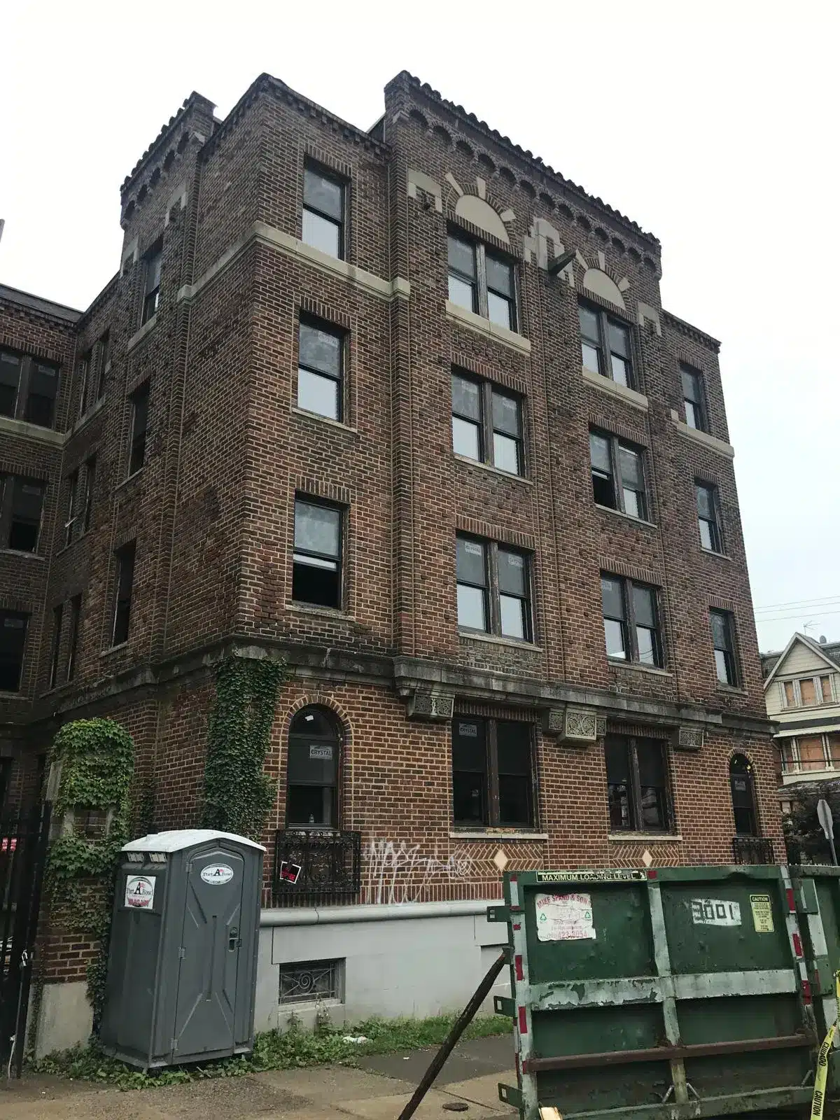Brick apartment building with graffiti, ivy on the lower wall, and a dumpster and portable toilet in front during renovation, relevant to rental insurance.